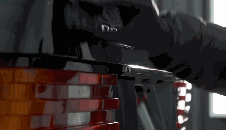 A person wearing black gloves opens the rear hatch of a classic car, revealing the taillight and glossy black exterior.