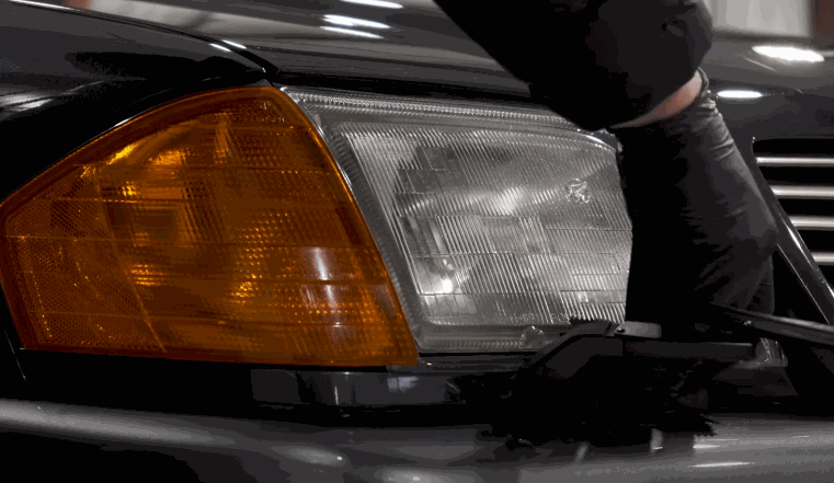 A person in black gloves gently applies ceramic coating to a vintage car, focusing on the headlight and front turn signal area.