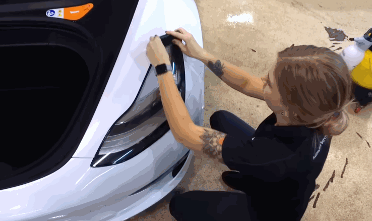 A technician installs paint protection film around the headlight of a white car, carefully pressing the material along the edge for full coverage.