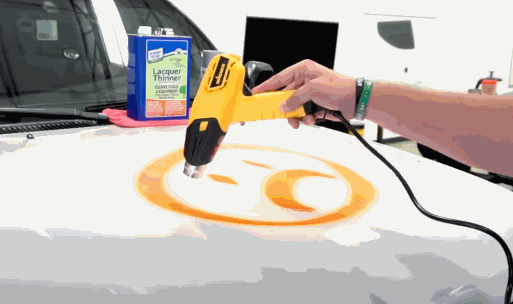 A heat gun is used to loosen a decal from a white vehicle hood during surface prep for paint protection film installation. A can of lacquer thinner is nearby.