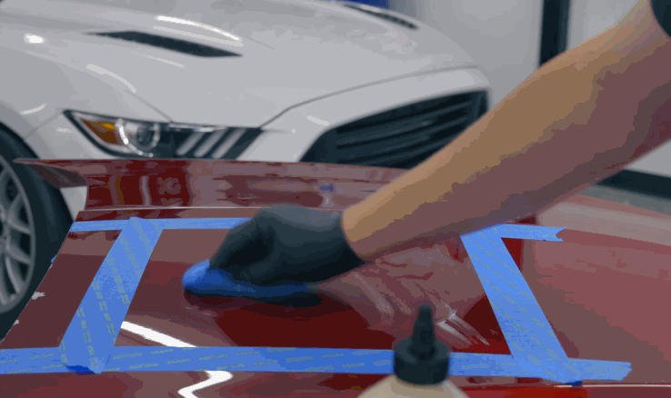 A gloved hand uses a blue applicator to wipe polish on a masked-off section of a red hood, refining the paint before PPF application.