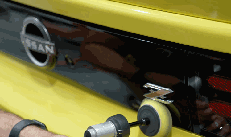 A rotary buffer is used to polish the black trim area on the rear of a yellow Nissan Z, likely as final prep before paint protection film.