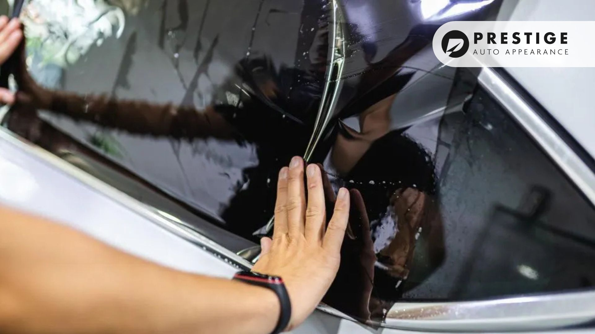 Hands pressing and smoothing window tinting film onto a car’s rear side window.