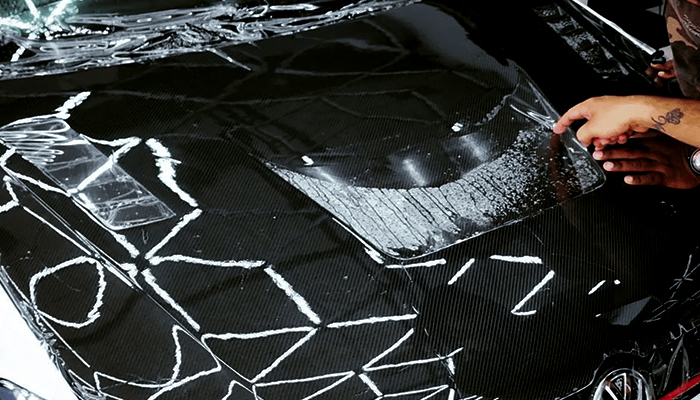 A technician presses down paint protection film onto the hood of a black car with a carbon fiber texture. The film has visible guide marks and water beneath for alignment.