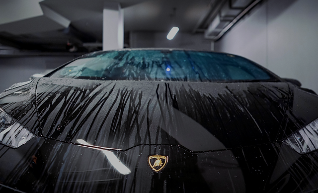 Water beads up and runs down the hood of a black Lamborghini parked indoors, showing the effect of a ceramic coating.
