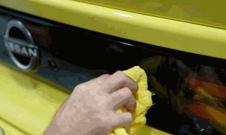 Close-up of a hand using a yellow microfiber cloth to polish the back of a yellow Nissan, focusing on the area near the Nissan logo.