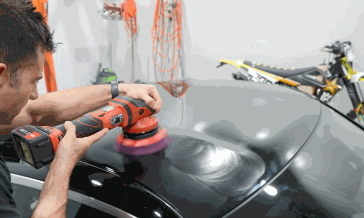A man uses a red dual-action polisher with a purple pad to buff the roof of a black car, reflecting garage lights and nearby equipment.