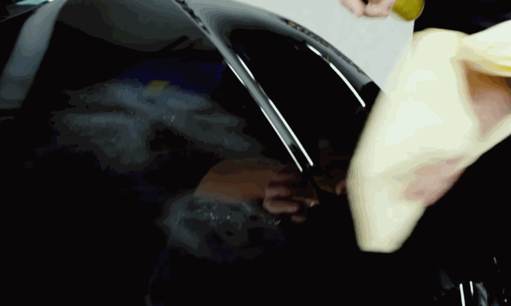 A person wipes down the side of a black car with a beige microfiber towel, removing polishing residue for a high-gloss finish.