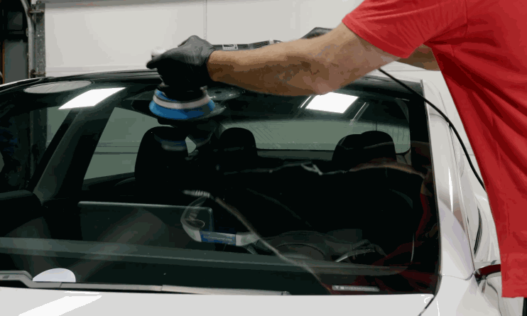A person in a red shirt and black gloves uses a dual-action polisher to clean the front windshield of a white car inside a garage.