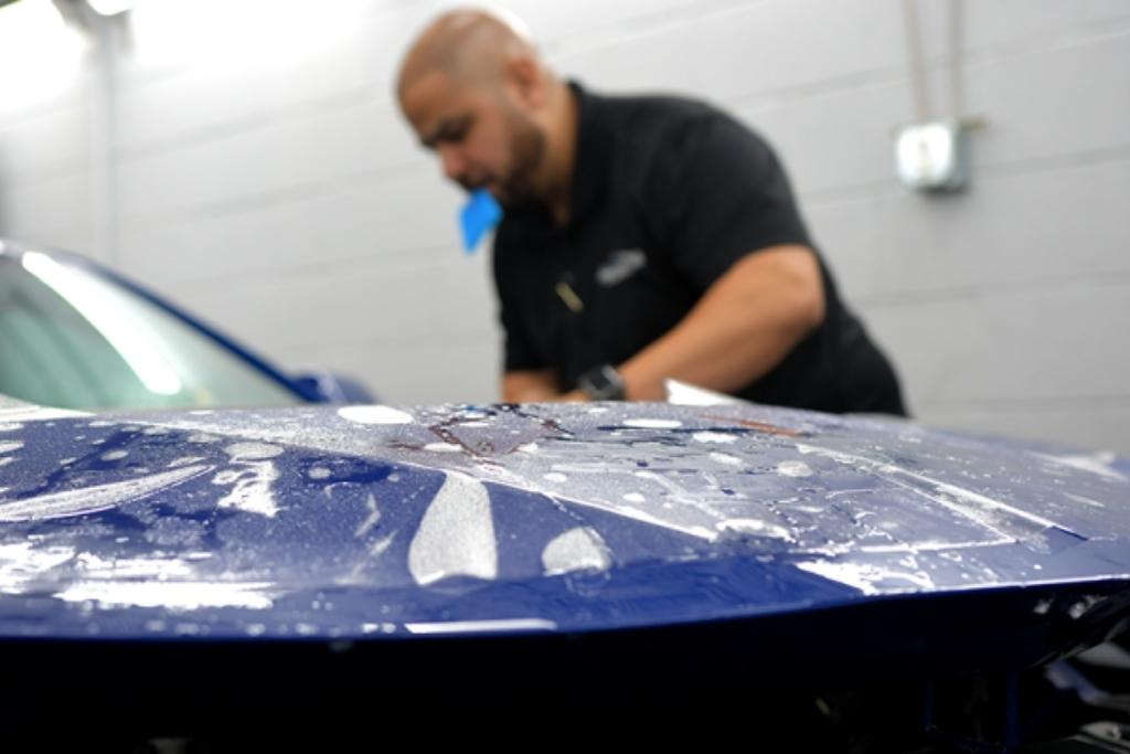 Auto detailing technician applying paint protection film to a blue vehicle hood in a professional workshop.