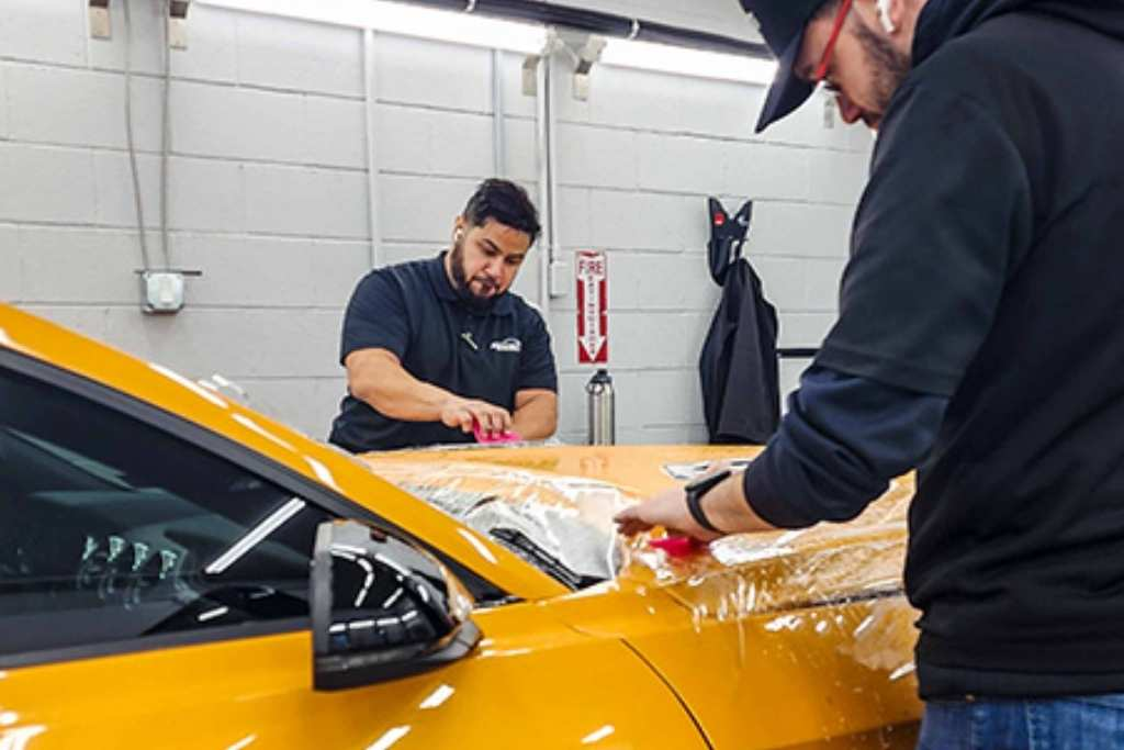 Two technicians applying paint protection film to the hood of a yellow car at a professional auto detailing studio.