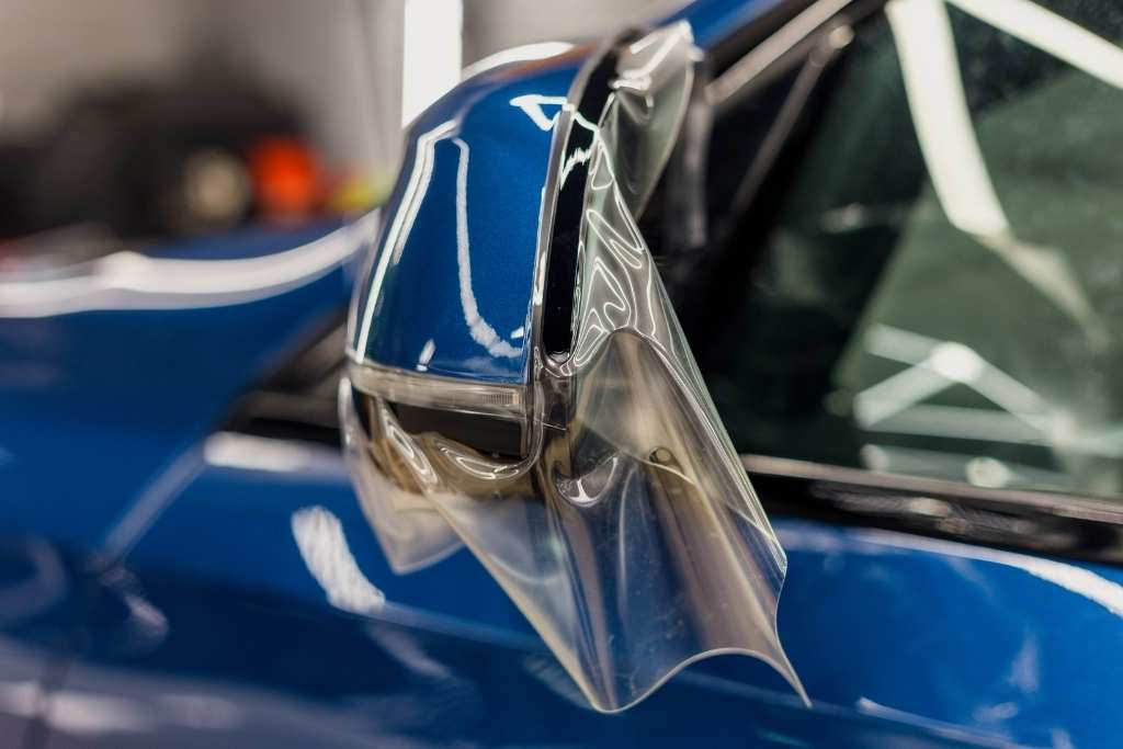 Clear paint protection film being applied to a blue vehicle's side mirror for added defense against chips and weather damage.