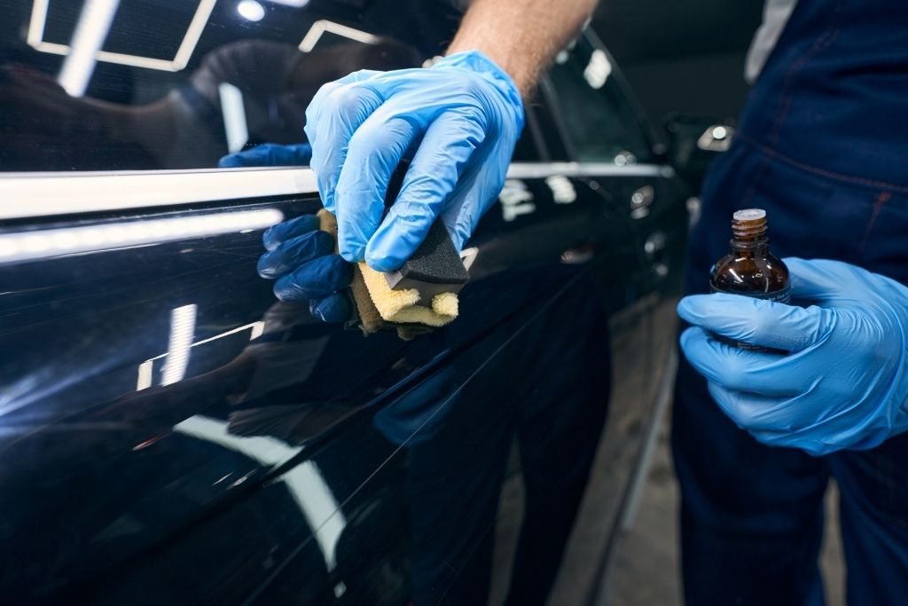 Detailing technician wearing blue gloves applying ceramic coating to a black vehicle door panel using an applicator block inside a professional auto detailing studio.