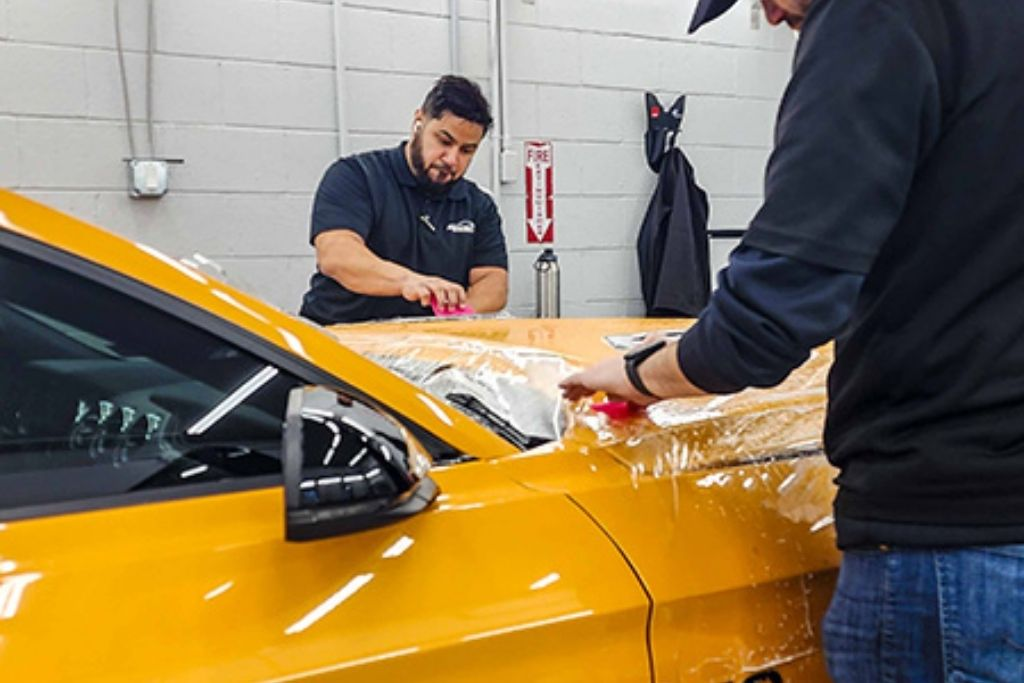 Technicians applying paint protection film to a yellow sports car hood inside a professional detailing studio.