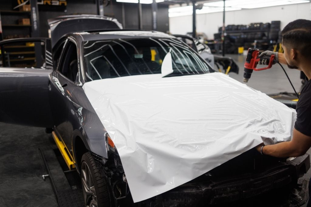 Technician using a heat gun to apply white vinyl wrap to the hood of a dark-colored car inside an auto shop.