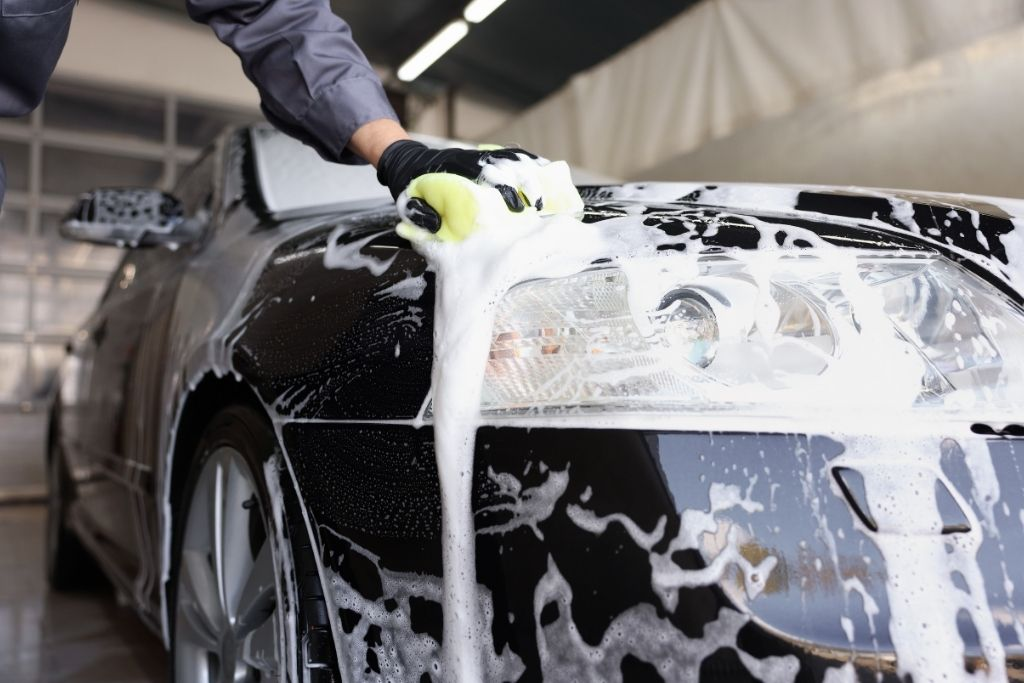 Person washing a black car with foam and sponge during professional exterior auto detailing.