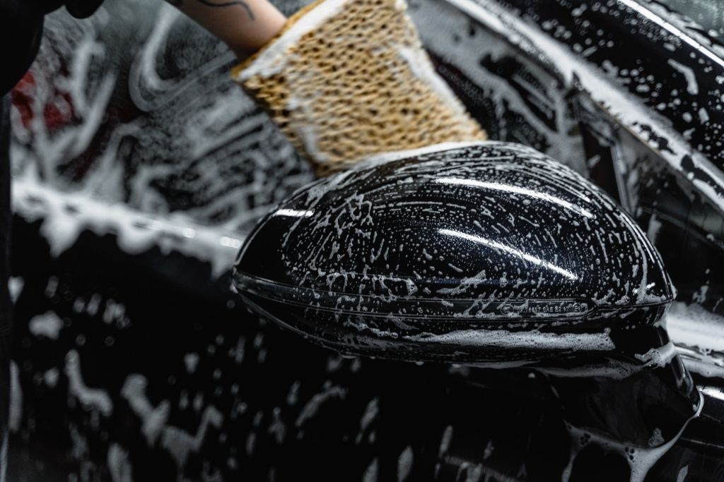 Detailing technician washing a black car side mirror with a sponge during professional exterior car detailing.