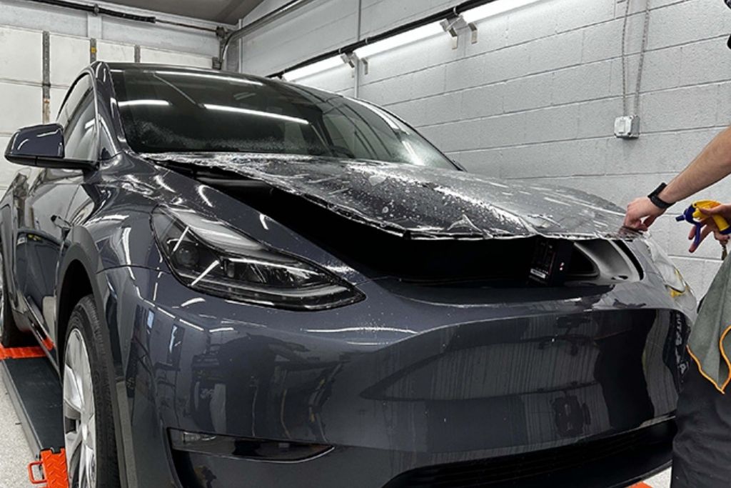 Installer applying paint protection film to the hood of a gray Tesla inside a professional detailing studio.