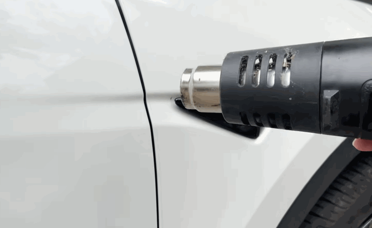 A heat gun warms the edge of paint protection film near a white vehicle door seam.