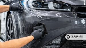 A technician smooths paint protection film across a gray front bumper below the headlight.