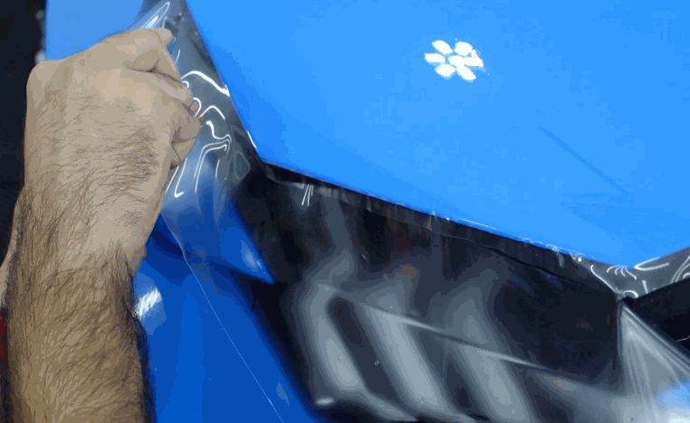 A technician pulls paint protection film around the headlight edge of a blue car during a close up installation.