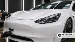 Technician applies paint protection film to the front bumper and headlight area of a white car.