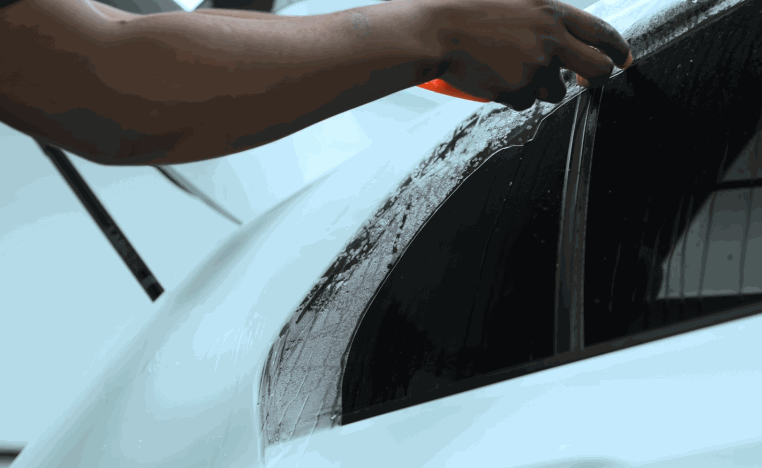 Installer pulls paint protection film into place over the rear quarter window area of a white car.