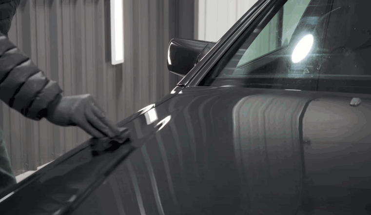 Detailer wearing a black glove wipes the gray body panel of a car under bright detailing lights to prepare the paint before ceramic coating application.