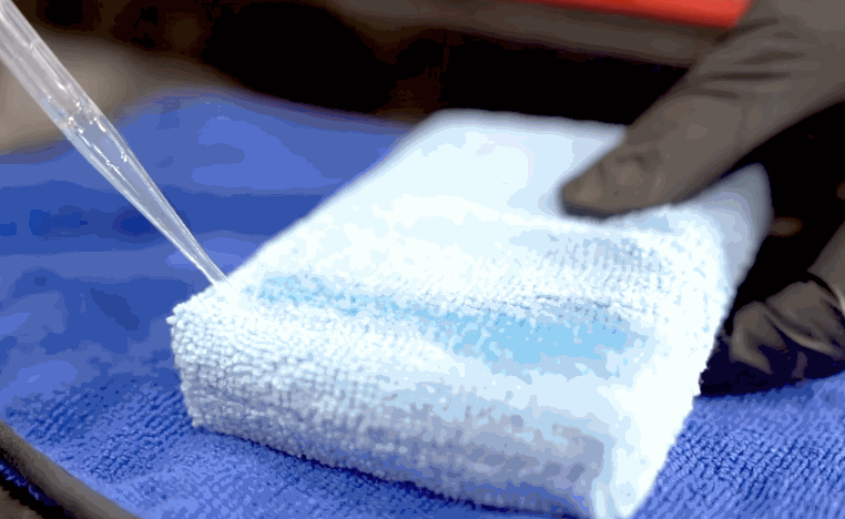 Ceramic coating liquid is dispensed from a dropper onto a white microfiber applicator pad resting on a blue towel.